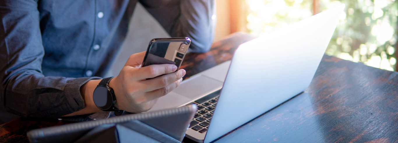 A man sat at his desk, with his computer, using his mobile phone.